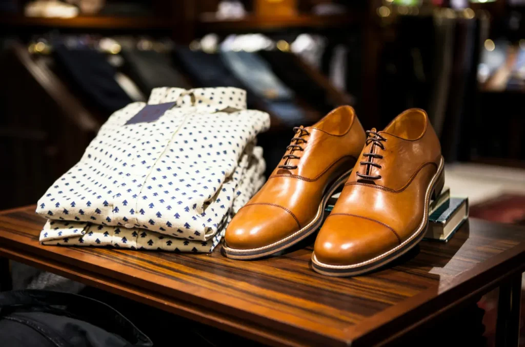 A folded patterned dress shirt and a pair of brown leather dress shoes are displayed on a wooden table in a clothing store.